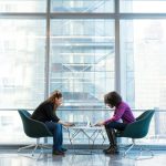 two women sits of padded chairs while using laptop computers